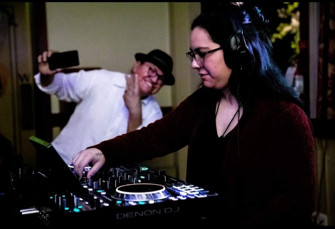 This candid, low-light photograph captures a woman actively engaged with DJ equipment in the foreground, while a man in the background playfully poses for a selfie. The woman is named Emily with long dark hair and wearing glasses and a dark red cardigan over a black top, is focused on the DJ mixer, her right hand adjusting a knob. The brand "DENON DJ" is visible on the equipment. She is also wearing over-ear headphones. Behind her, slightly out of focus, a man who is named Marco is wearing a white shirt and a dark fedora is holding up a smartphone, seemingly taking a selfie. He is smiling broadly and making a peace sign with his left hand. The lighting suggests an indoor setting, possibly a party or event, with some ambient light illuminating the subjects.