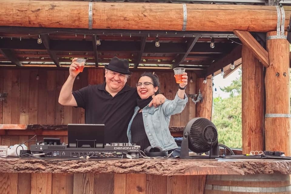 This photograph shows two people, a man, Marco and a woman, Emily standing behind a rustic wooden bar with a  DJ booth, raising their drinks in a celebratory gesture.  The man, on the left, is wearing a black polo shirt and a dark fedora. He has a wide smile and is holding up a clear plastic cup with an orange beverage. His left arm is around the woman's shoulders.  The woman, on the right, is wearing glasses, a black shirt, and a light blue denim jacket. She also has a wide smile and is holding up a clear plastic cup with an orange beverage. In front of them on the wooden counter, there's a laptop, DJ equipment (with "DENON DJ" visible), headphones, and a black fan. The structure they are in appears to be made of logs and wood, with exposed beams and hanging lights overhead, suggesting an outdoor or semi-outdoor venue with a natural, rustic aesthetic.