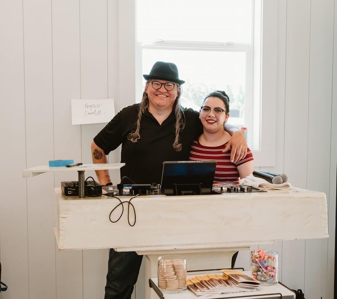 This indoor photograph shows two people, a man and a woman, ( Marco and Emily)  standing behind a white DJ booth or table, both smiling at the camera.  The man, on the left, has long, wavy dark hair, glasses, and a black fedora. He is wearing a black collared shirt and dark pants, and his right arm is around the woman's shoulder. A tattoo is visible on his left forearm.  The woman, on the right, has dark hair pulled back, glasses, and is wearing a red and white striped t-shirt. She is also smiling.  On the white table in front of them, there's DJ equipment, a laptop, and microphones. To the far left, a piece of white paper with "Marco + Emily" written in cursive is taped to a stand. Below the table, a clear jar filled with round, light-colored objects (possibly cookies or crackers) and some brochures are visible. Another clear container filled with colorful candies is also on a lower shelf to the right. The background features a white wall with vertical paneling and a large window, letting in natural light.