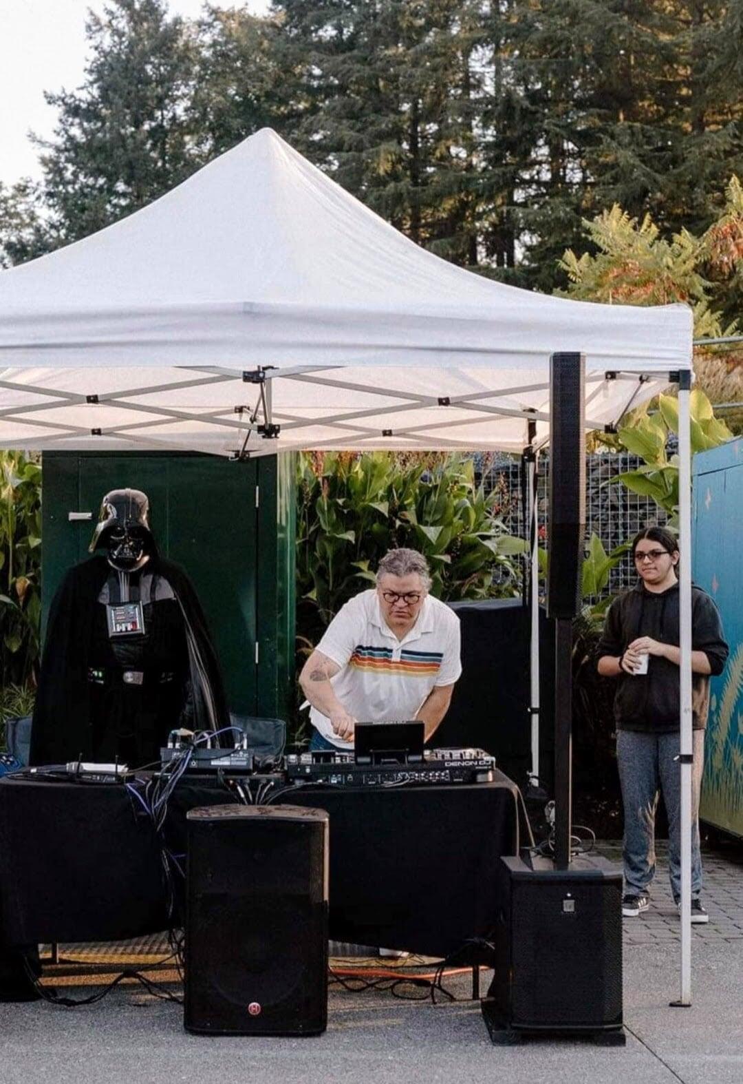 This photo captures an outdoor DJ setup under a white canopy, featuring three main figures and various equipment.  In the center, under the canopy, a man, Marco,  with glasses and a light-colored polo shirt with horizontal stripes is actively DJing, his hands on a mixer. To his left, a person dressed as Darth Vader stands beside the DJ booth. On the right side of the frame, partially under the canopy's edge, a younger individual, Ian,  with dark hair and glasses is standing, holding a cup.  The DJ booth is set up with black tablecloths, and there are speakers on the ground in front of and to the side of the booth. The background shows lush green foliage and trees, indicating an outdoor event. The lighting suggests it's either late afternoon or early evening.