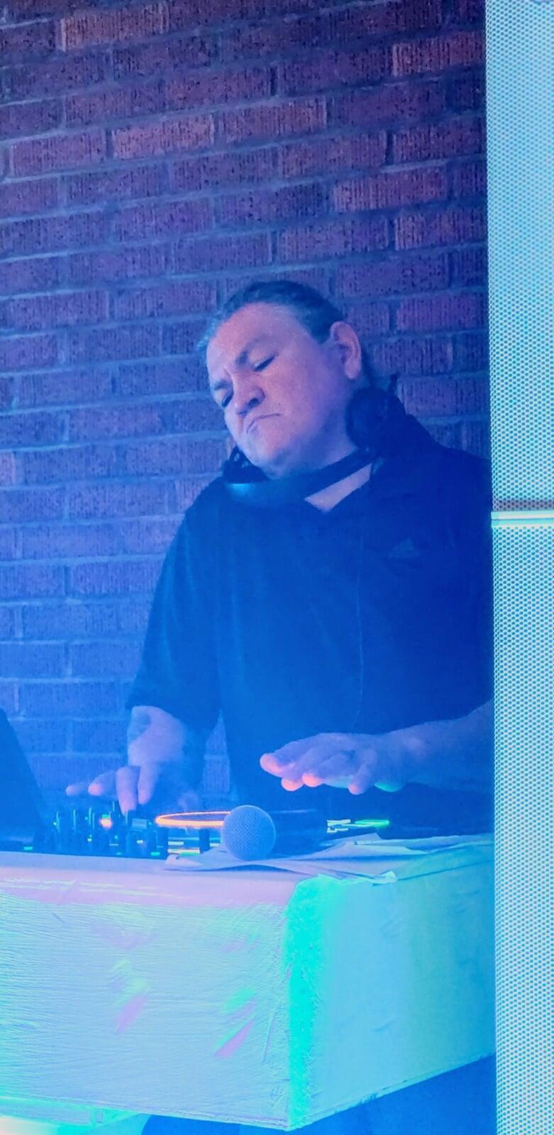 This full-body, portrait-style shot shows a man, Marco , standing behind a DJ setup. He appears to be actively mixing music, with his hands positioned over a DJ controller. He is wearing headphones around his neck and a dark t-shirt, possibly black or a dark navy, with a faint Adidas logo visible on the right side of his chest.  The background is a brick wall, illuminated by purple and blue lights, creating a vibrant, club-like atmosphere. In the foreground, the DJ booth itself is lit with a cool, bluish-white light, and a microphone is visible on the table. To the right of the frame, part of a white speaker with a perforated pattern is visible. The man has darker hair and is looking down at his equipment, seemingly focused on his task.